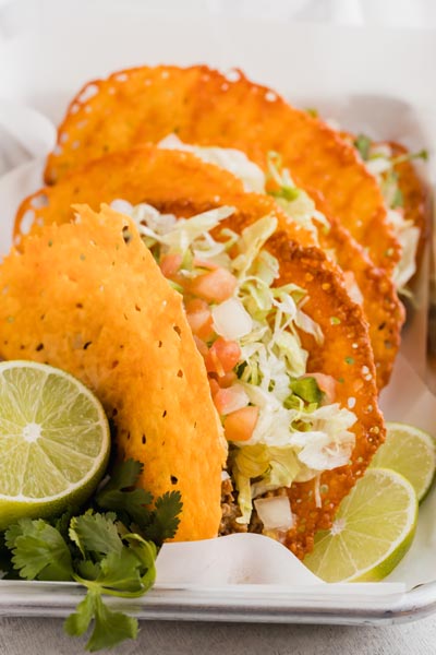 Crispy hard shell taco made out of cheese and filled with beef, lettuce and pico de gallo next to cilantro and limes.