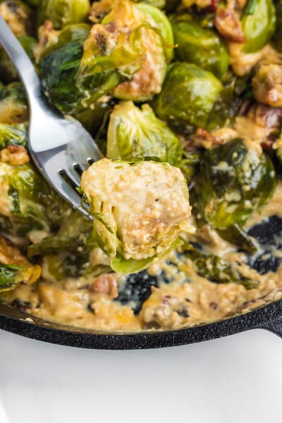 A fork holding a creamy brussel sprout in front of the skillet with more.