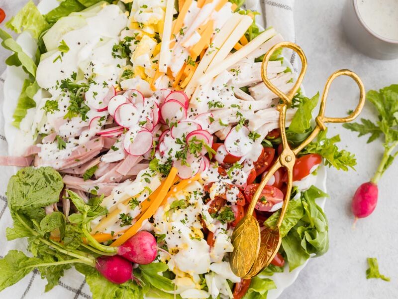 Looking down onto a chef salad topped with ranch dressing, fresh parsley and tongs nearby.