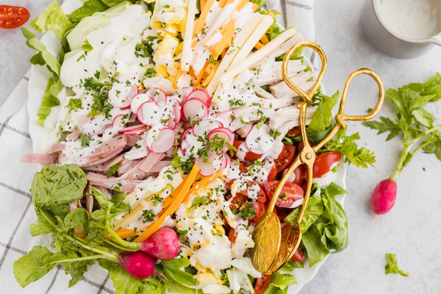 Looking down onto a chef salad topped with ranch dressing, fresh parsley and tongs nearby.