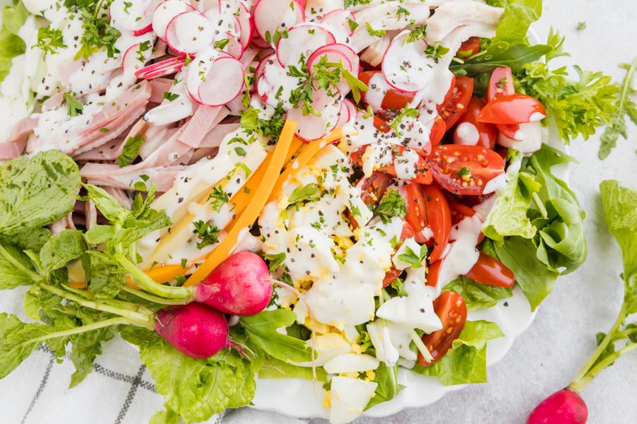 A large chef salad on a plate filled with tomato, egg, cheese, turkey and radishes. Topped with ranch dressing.