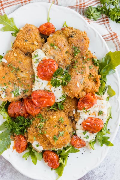 A platter with breaded chicken, tomatoes and melted cheese on top of arugula.