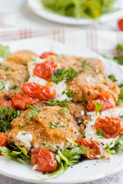 Crispy breaded chicken on a platter with tomatoes, melted burrata cheese and parsley.