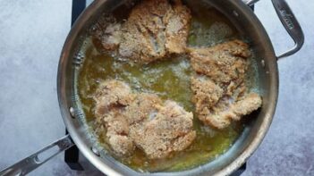 A frying pan with three crusted chicken thighs cooking. Oil bubbles around.
