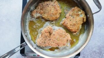 Three chicken thighs coated with pork panko frying in a large stainless steel skillet.