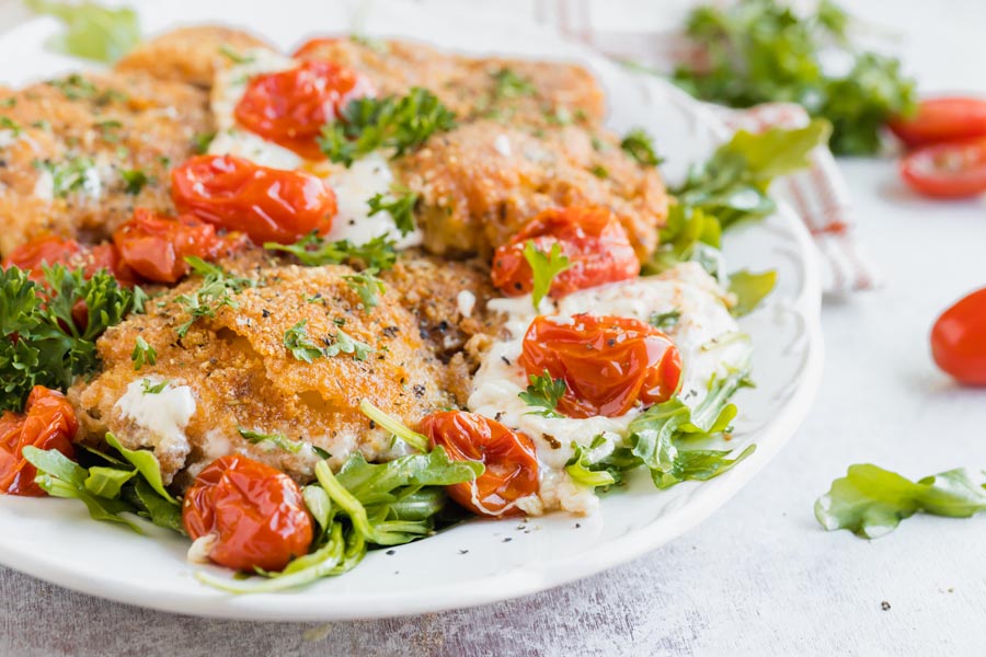 A platter of breaded chicken with melted burrata cheese, sauteed tomatoes and arugula.