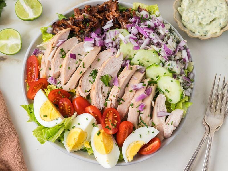 Overhead view of a loaded chicken cobb salad with sliced chicken breast, bacon, cucumbers, eggs, blue cheese, and red onion.