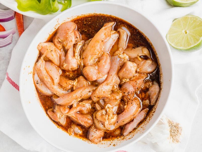 Sliced chicken thighs marinade in a bowl with slice of lime nearby.