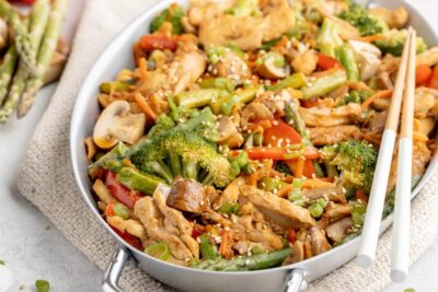 a skillet with a colorful mix of veggies and stir fry chicken topped with sesame seeds with chopsticks hanging over