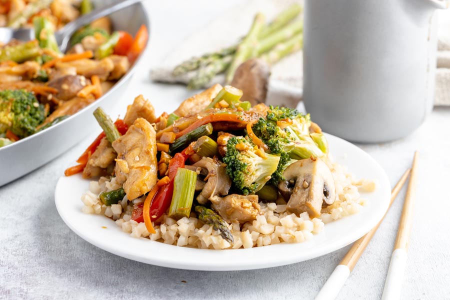 a small dinner plate with chicken and vegetable stir fry over rice next to chopsticks and skillet with more stir fry