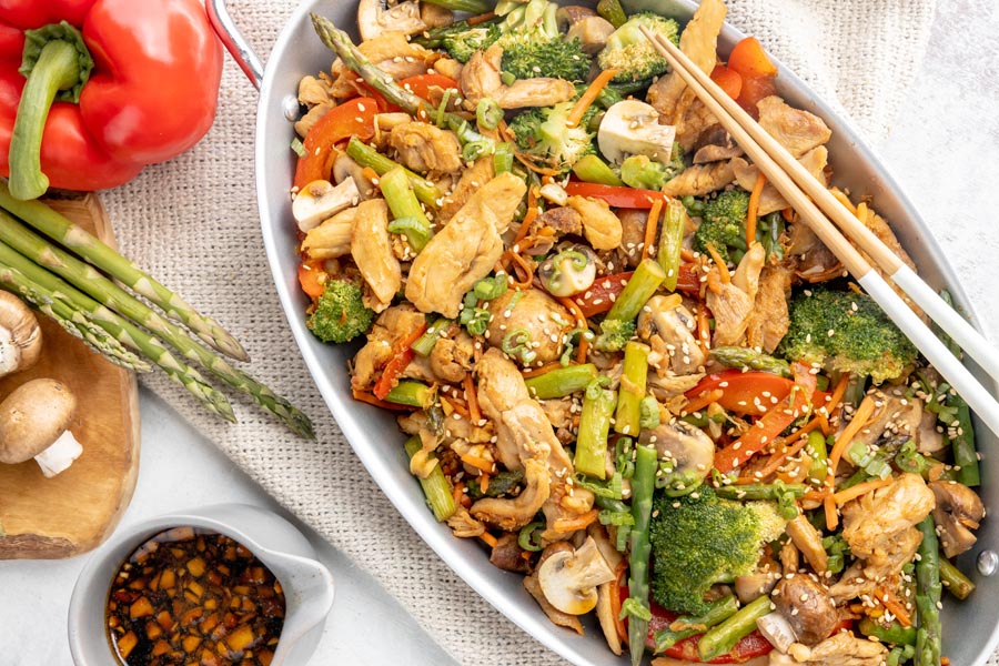 a skillet with homemade veggie and chicken stir fry next to fresh asparagus, red bell pepper, mushrooms and more stir fry sauce