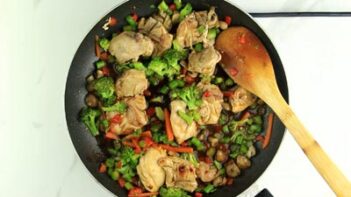 a wooden spoon stirs a skillet with chicken, vegetables and stir fry sauce