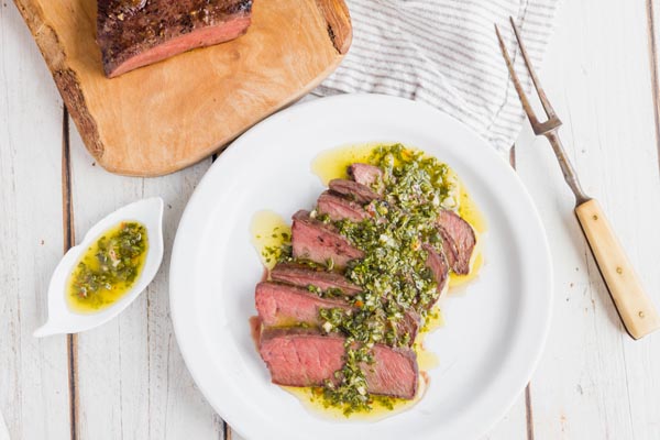 a plate of steak covered in chimichurri sauce with a fork and more steak nearby