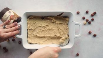 spreading cake batter into a baking dish with hazelnuts scattered around