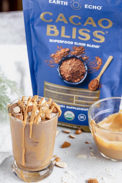 a blue bag of cacao bliss sits behind a small chocolate smoothie topped with nuts and a bowl of peanut butter next to it