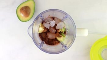 a blender with avocado, ice and cocoa powder inside with a half an avocado next to it