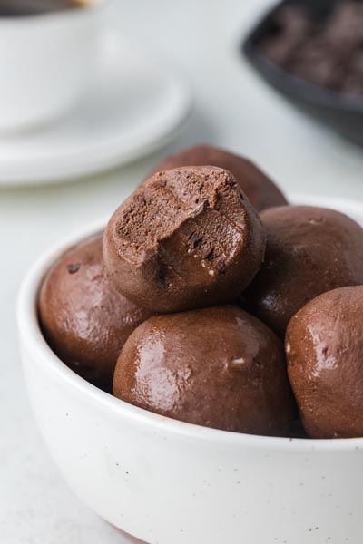 A bowl of chocolate protein balls, one with a bite revealing the smooth, fudgy interior.