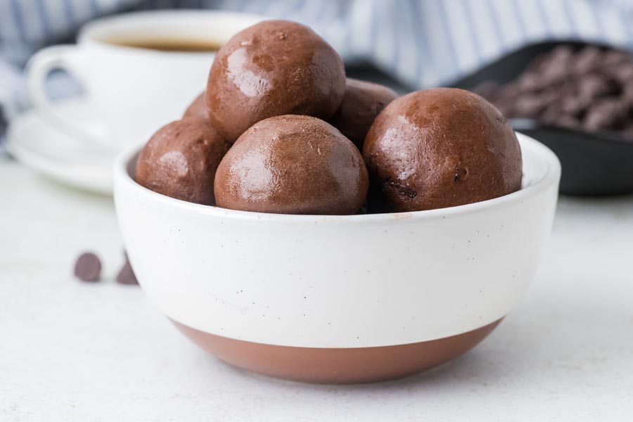 A white and tan bowl holding several glossy chocolate protein balls on a light background.