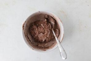 A mixing bowl with chocolate protein ball dough and a spoon on a light surface.
