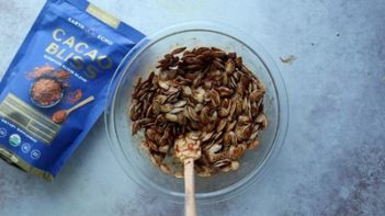 chocolate coated pumpkin seeds in a bowl next to a bag of cacao bliss