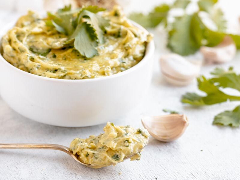 A spoonful of creamy cilantro aioli in front of garlic, cilantro leaves and a bowl of aioli.