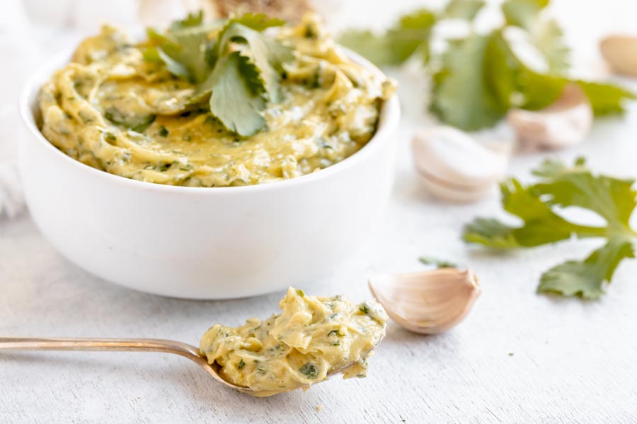 A spoonful of creamy cilantro aioli in front of garlic, cilantro leaves and a bowl of aioli.