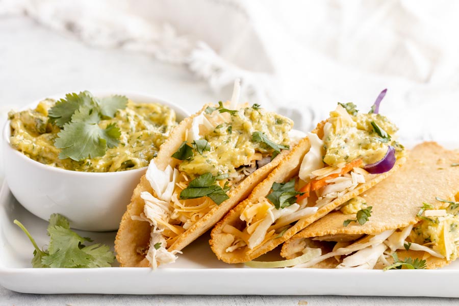 Three tacos topped with cabbage and cilantro lime aioli on a rectangle plate with a white bowl of more aioli in the background.