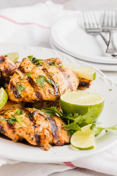 A plate of grilled chicken thighs with lime as a garnish and two plates in the background with forks.