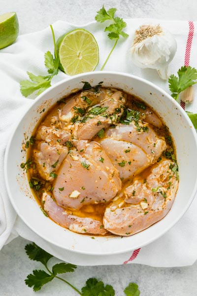 A bowl with raw chicken marinating in a cilantro lime mixture with garlic and limes nearby.