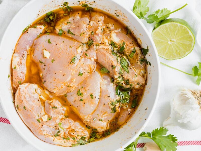 A bowl of chicken thighs marinating with a half a lime, garlic and cilantro nearby.