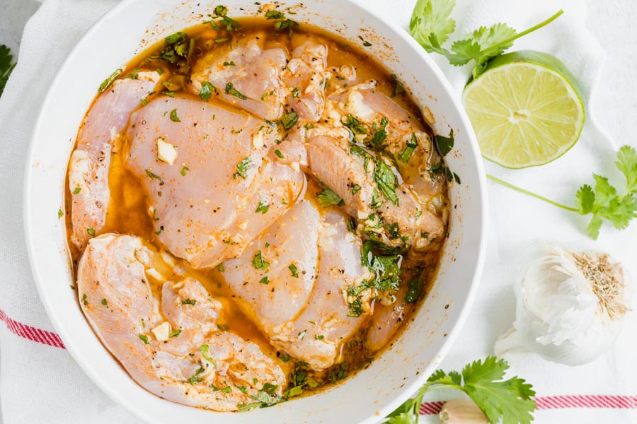 A bowl of chicken thighs marinating with a half a lime, garlic and cilantro nearby.