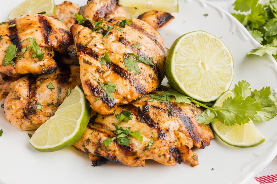 A pile of grilled chicken thighs on a platter with limes and cilantro all over.