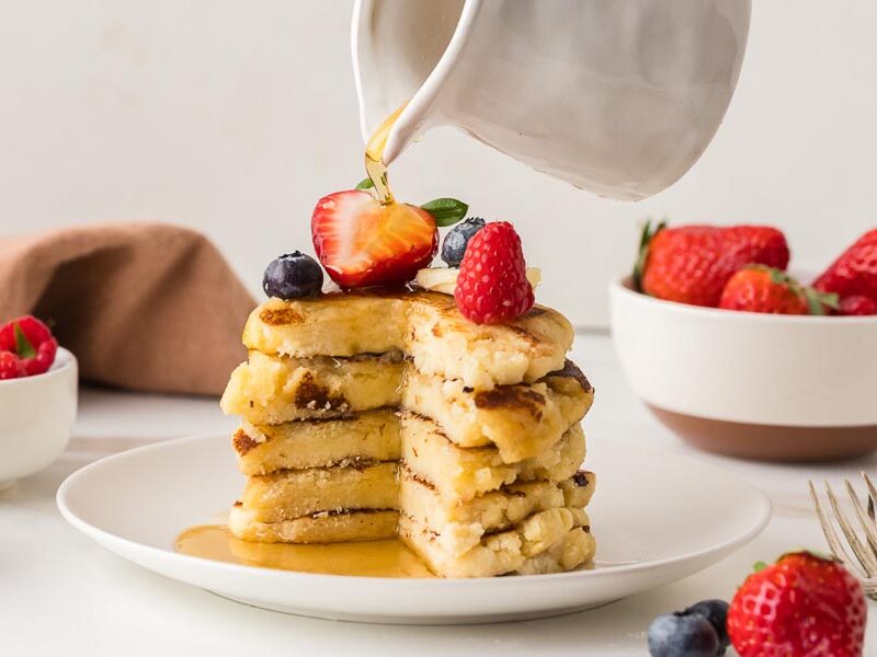 Stack of cottage cheese pancakes with berries as maple syrup pours from a pitcher.