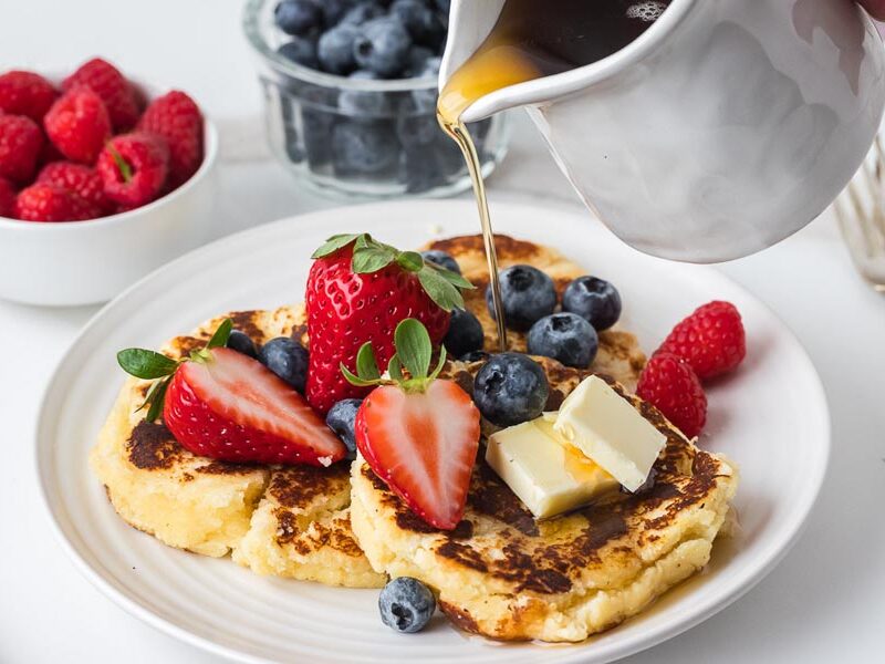 Golden cottage cheese pancakes topped with butter, fresh strawberries, blueberries, raspberries, and a drizzle of sugar-free maple syrup.