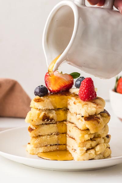 Sugar-free maple syrup being poured over a stack of fluffy cottage cheese pancakes topped with fresh berries and butter.