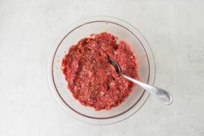 Raw ground beef mixed with seasonings, herbs, and other meatball ingredients in a glass mixing bowl.
