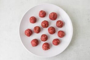 Twelve evenly sized raw meatballs rolled and arranged on a white plate before cooking.