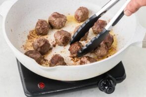 Homemade meatballs being browned in a skillet with cooking tongs until golden on all sides.