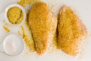two chicken breasts on a cutting board covered with lemon pepper plus two dishes of salt and lemon pepper nearby
