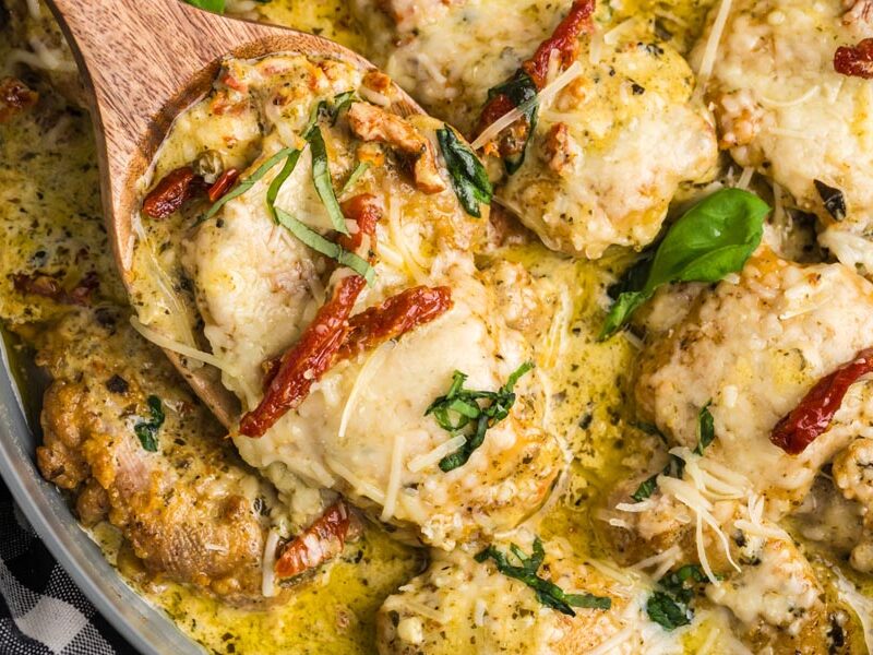 A slice of pesto chicken on a wooden spoon topped with basil and sun-dried tomatoes.