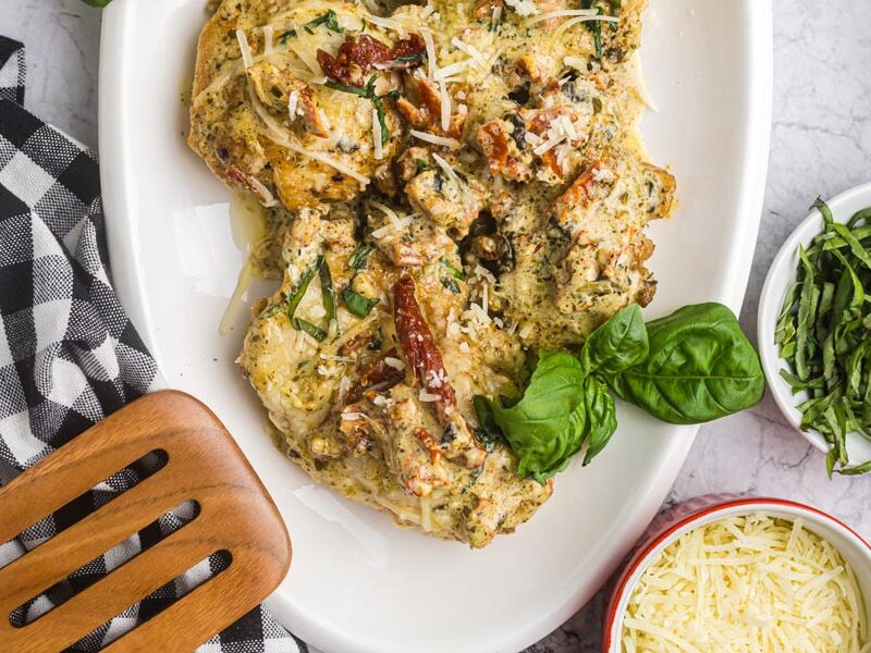 Pesto chicken on a serving platter next to sliced basil and shredded parmesan cheese.