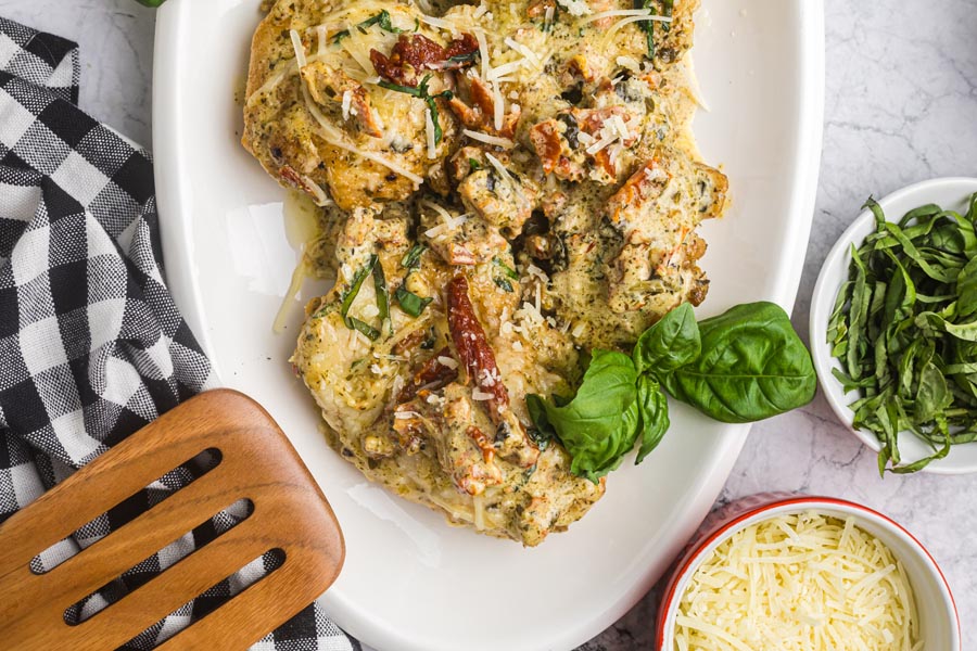 Pesto chicken on a serving platter next to sliced basil and shredded parmesan cheese.