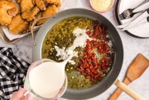 Pouring heavy cream in a skillet with pesto and sun-dried tomatoes.