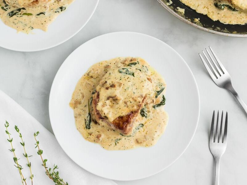 A smothered pork chop on a white plate topped with a creamy Dijon bacon gravy. With forks and fresh thyme nearby.