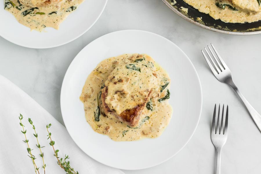 A smothered pork chop on a white plate topped with a creamy Dijon bacon gravy. With forks and fresh thyme nearby.