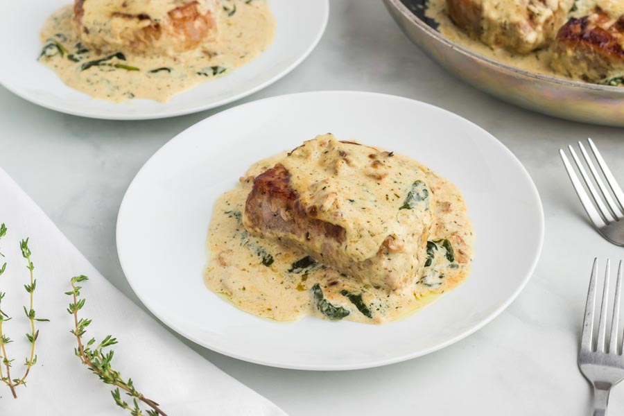 A creamy spinach sauce covered a boneless pork chop on a white plate with another plate behind and two forks to the right.