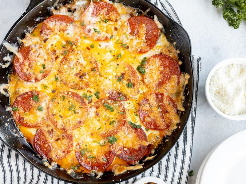 looking down on a cast iron skillet with a pepperoni pizza inside while a bowl of parmesan cheese sits next to it