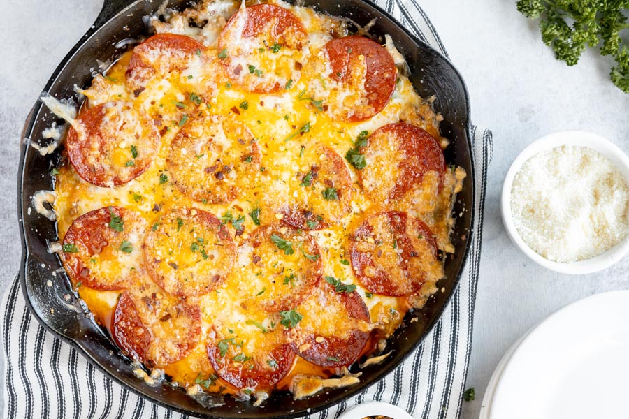 looking down on a cast iron skillet with a pepperoni pizza inside while a bowl of parmesan cheese sits next to it