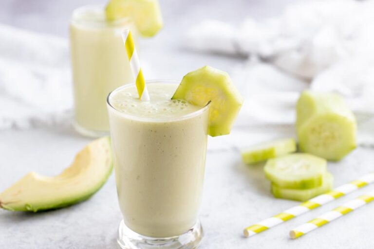 a smoothie made with avocado and cucumber sits in front of slice cucumbers with a striped yellow straw in it