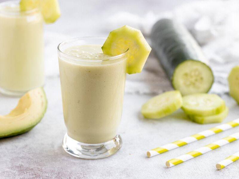 a green cucumber smoothie sits in front of a sliced cucumber, avocado and straws